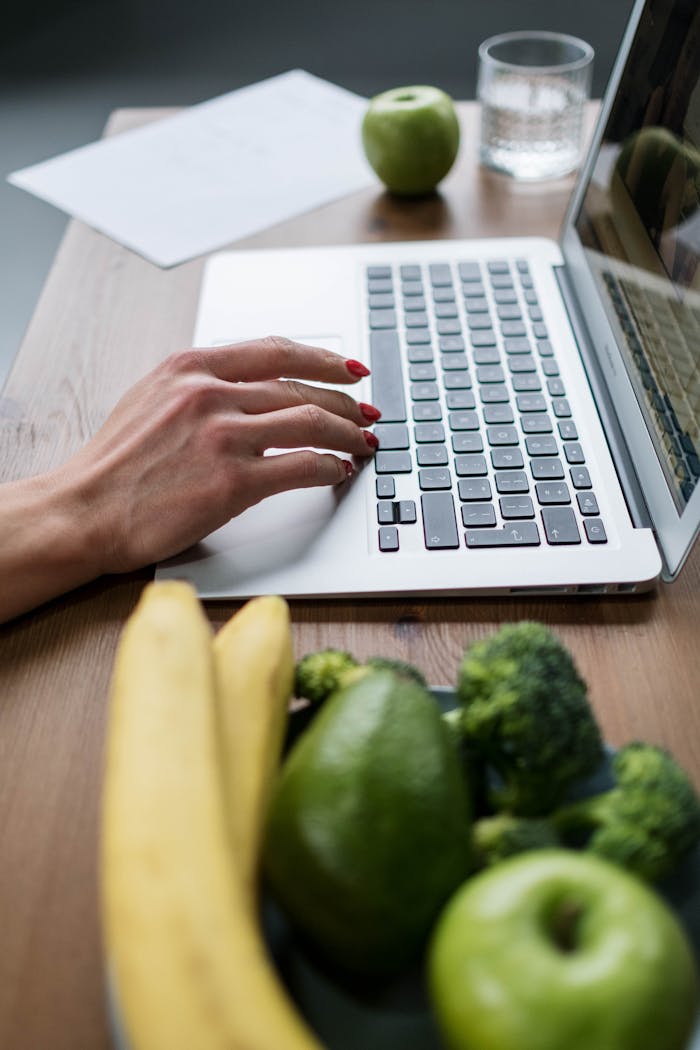 Close-up of a womans hand with red nails typing on a laptop, with fresh fruits and vegetables on the table.