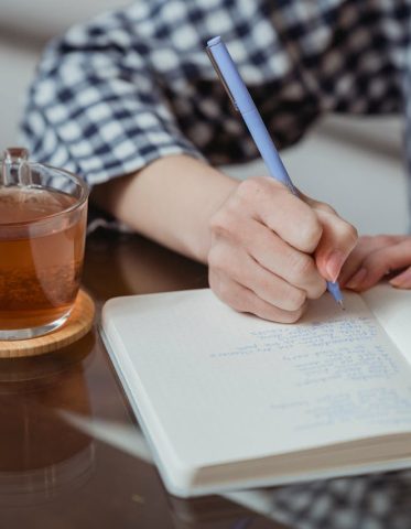 Close-up of hands writing in a notebook beside a glass of tea on a wooden coaster.